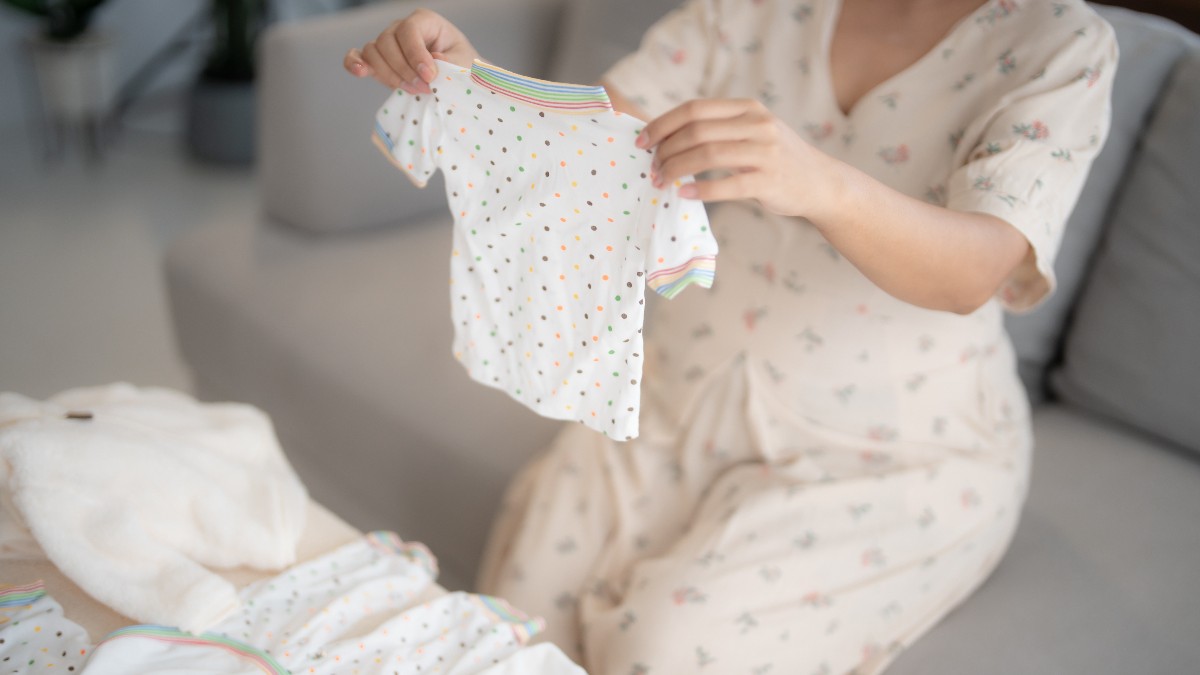 Imaginea surprinde o mamă care pregătește haine pentru bebeluș, ținând în mâini un body alb pentru copii. Fotografia ilustrează grija pentru alegerea hainelor celor mici și se potrivește unui articol despre închirierea articolelor vestimentare pentru bebeluși și copii.