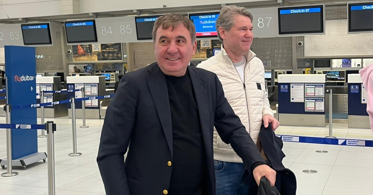Gică Hagi în aeroport într-un costum negru, în timp ce zâmbește.