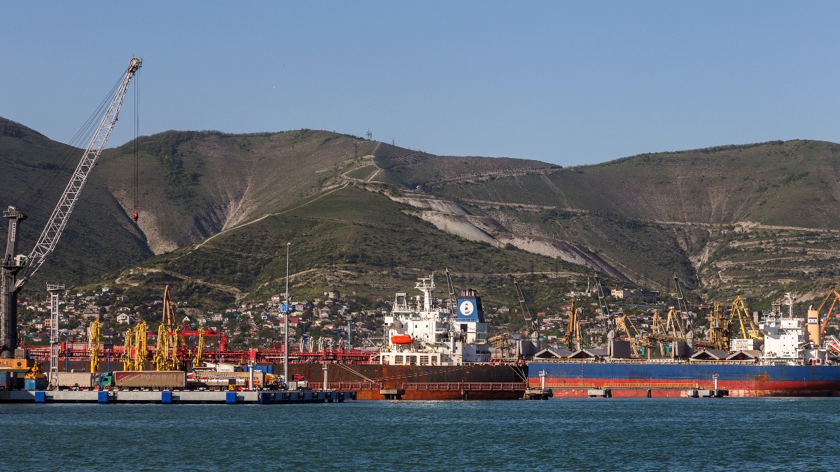 În imagine se vede un port maritim, cu mai multe nave comerciale și instalații pentru încărcarea și descărcarea mărfurilor. În fundal se află un relief muntos, iar zona portuară este dominată de macarale și infrastructură industrială.