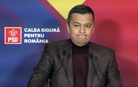 Sorin Grindeanu, îmbrăcat într-o maletă vișinie și un sacou maro cu model de caroruri gri, făcând o grimasă, cu buzele strânse și încovoiate în jos, și ochii ațintiți spre stânga