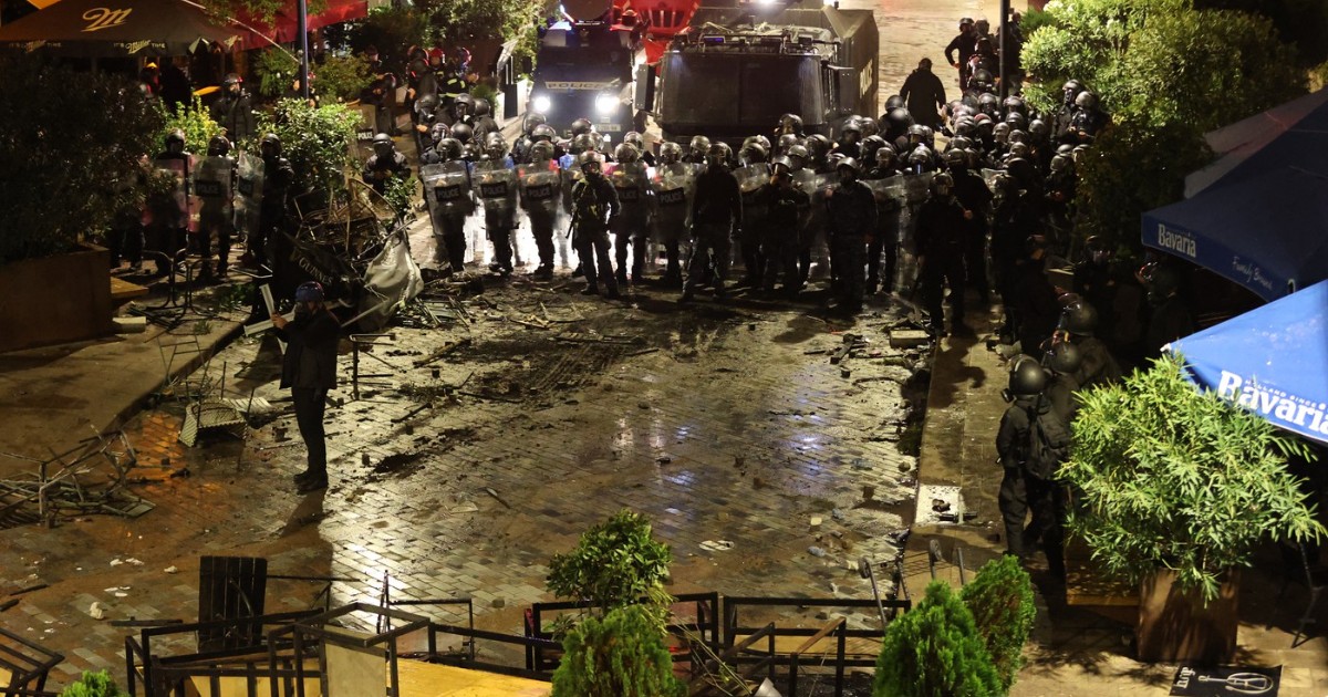 Imaginea surprinde o scenă de tensiune nocturnă, unde un rând compact de forțe de ordine (posibil poliție sau jandarmerie) în echipament de intervenție se confruntă cu o zonă dezordonată și plină de resturi. În prim-plan se văd mese și scaune răsturnate, iar pe fundal, două vehicule blindate și un grup mare de agenți menținând o linie de apărare, sugerând o intervenție după tulburări civile sau un protest.