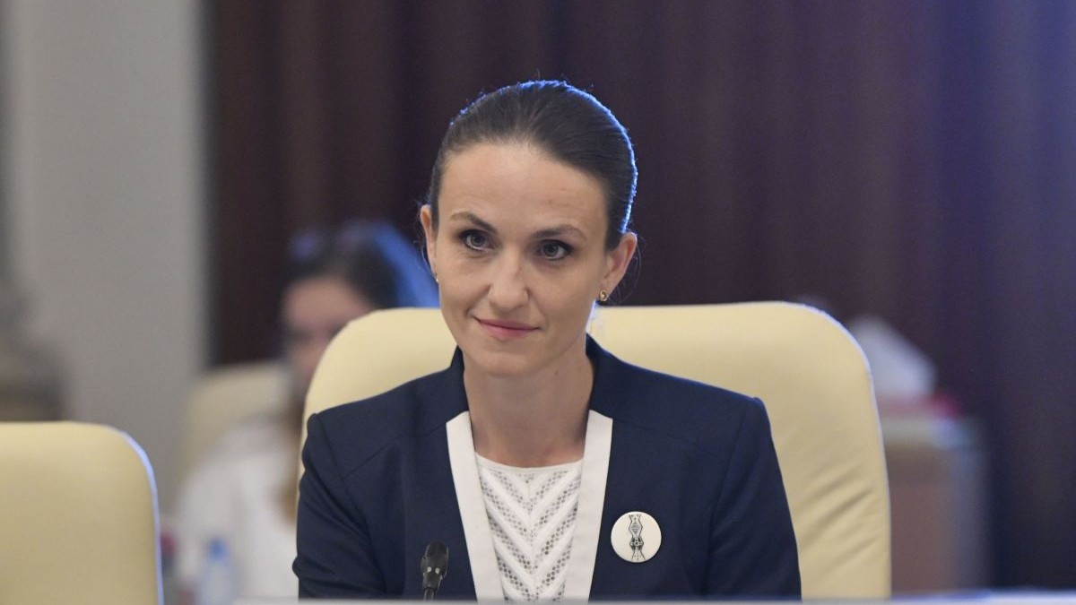 Oana Țoiu, îmbrăcată cu o bluză albă și un sacou albastru, în timp ce stă pe un scaun de culoare crem