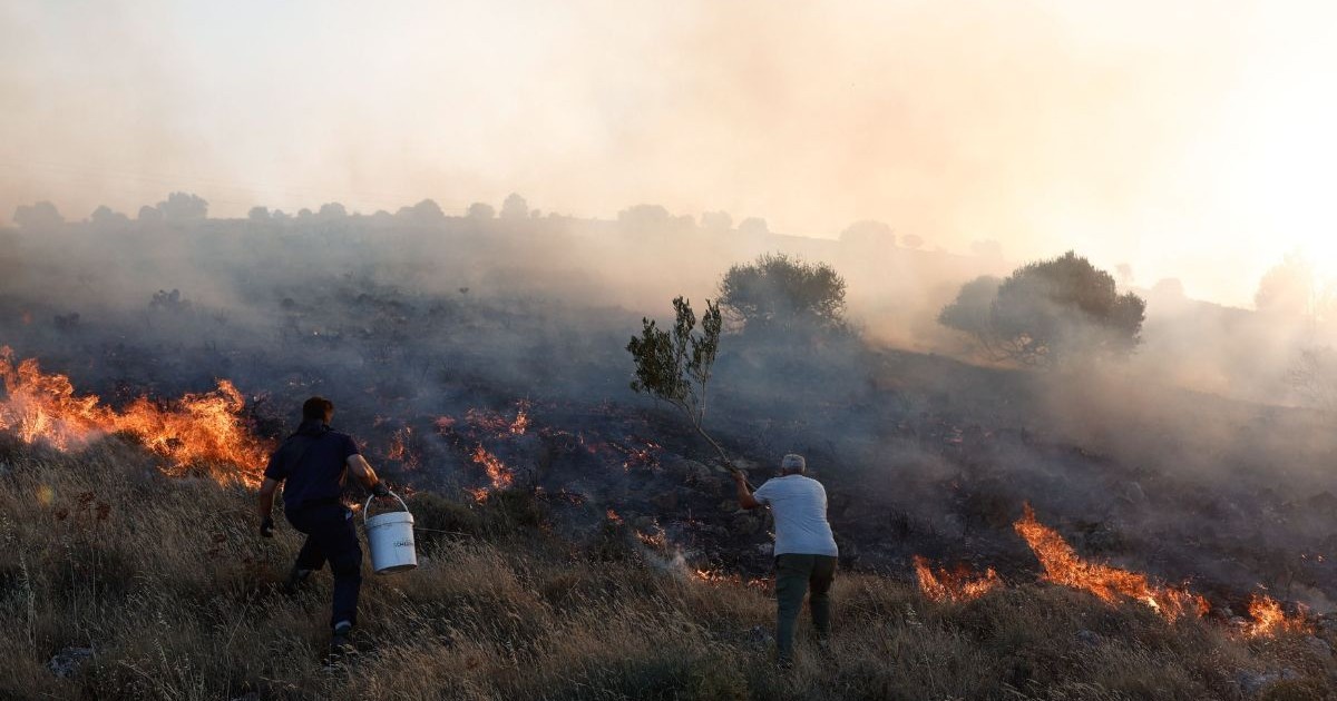 pajiște cu iarbă uscată în flăcări, Chios, Grecia, 22 iunie. fum gros de ridică din incendiul de vegetație. doi bărbați ,cu spatele spre cameră, încearcă să stingă flăcările cu găleți cu apă albe. Bărbatul din stânga este îmbrăcat într-o pereche de pantaloni negri și un tricou negru iar bărbatul din dreapta poartă o pereche de pantaloni negri și un tricou alb