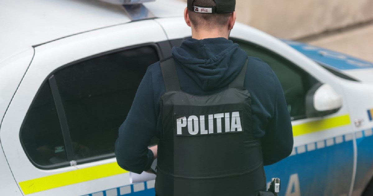 Imaginea surprinde, din spate, un bărbat purtând o vestă antiglonț pe care scrie „POLIȚIA”, stând lângă o autospecială de poliție inscripționată.