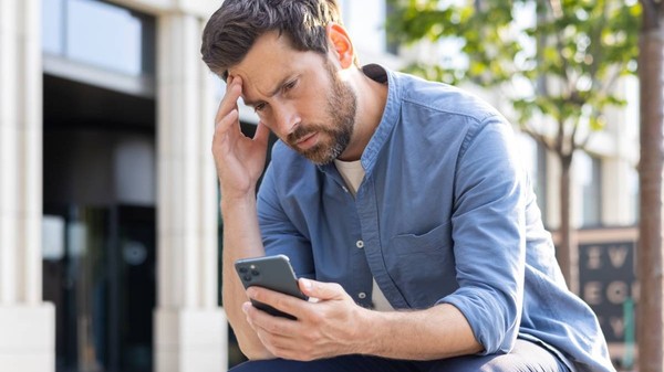 Un tânăr supărat stă pe o bancă pe o stradă a orașului și privește dezamăgit ecranul telefonului pe care îl ține în mână.