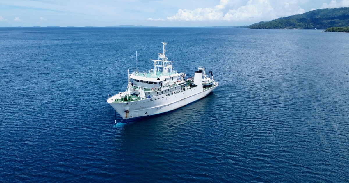 Imaginea prezintă o navă de cercetare maritimă care navighează pe o mare calmă, în larg, având în fundal o zonă de coastă cu relief împădurit. Nava este echipată cu instalații specifice misiunilor științifice și tehnice, sugerând activități de explorare sau exploatare a resurselor marine.