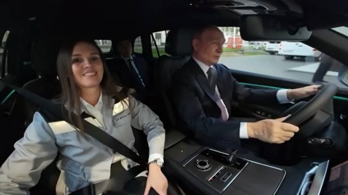 Vladimir Putin conduce un SUV Volga, îmbrăcat într-un costum închis la culoare și cravată. Pe scaunul din dreapta se află o femeie purtând centura de siguranță, iar în spate se vede un alt pasager. Imaginea este surprinsă din interiorul mașinii, în timp ce vehiculul se deplasează printr-o zonă industrială.