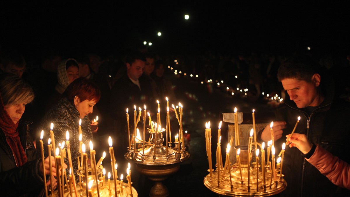 Mai multe persoane aprind lumânări într-un cadru religios, în timpul nopții, creând o atmosferă solemnă și liniștită. Flăcările lumânărilor luminează fețele credincioșilor, sugerând un moment de reculegere și rugăciune.