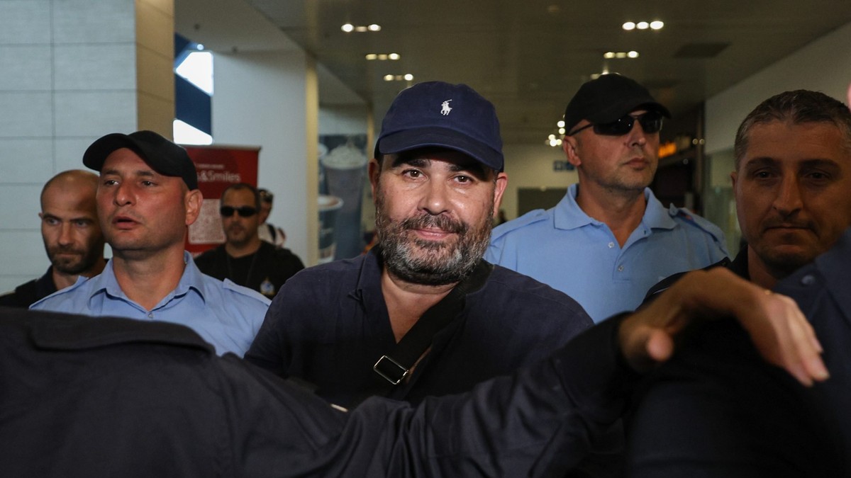 Imaginea îl arată pe Darius Vâlcov, cu barbă și șapcă, flancat de mai mulți polițiști.