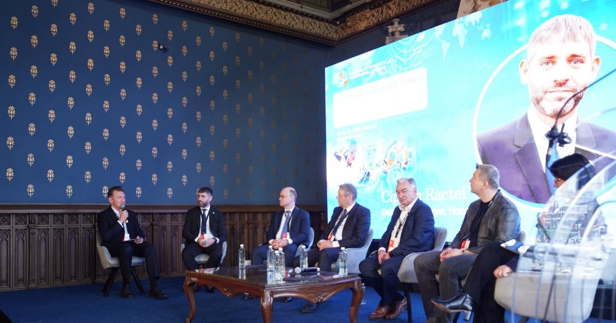 Mai mulți lideri din mediul de afaceri participă la un panel de discuții organizat într-o sală elegantă, în cadrul unei conferințe. Pe scenă, invitații discută despre teme economice, în timp ce în spate este proiectat un ecran cu informații despre eveniment.
