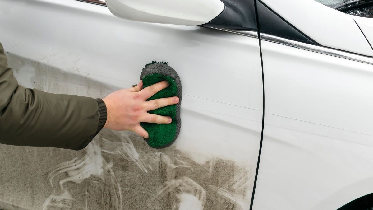 Mâna unui bărbat îmbrăcat cu o geacă închisă la culoare, care ține un burete negru, cu care șterge portiera stângă a unei mașini murdare, de culoare albă