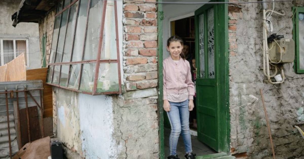 O fetiță stă în pragul unei case vechi, cu ușa verde deschisă. Clădirea are pereți deteriorați, cu tencuială căzută, cărămidă la vedere și ferestre vechi. În jur se văd improvizații, cabluri și obiecte sprijinite de pereți, într-un spațiu care sugerează condiții modeste de locuire. Imaginea transmite vulnerabilitatea copiilor care cresc în sărăcie.