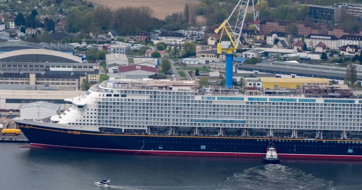 Nava de croazieră ancorată în marină, pe apă, macarale si un oraș în fundal, alte bărci mici pe lângă