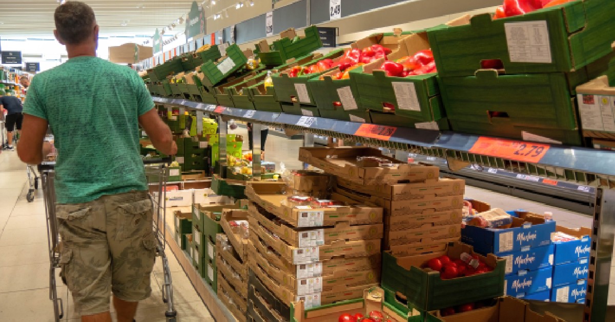 Imaginea surprinde un client împingând un cărucior de cumpărături pe culoarul unui supermarket, trecând pe lângă rafturi pline cu lăzi de legume proaspete, precum ardei și roșii.