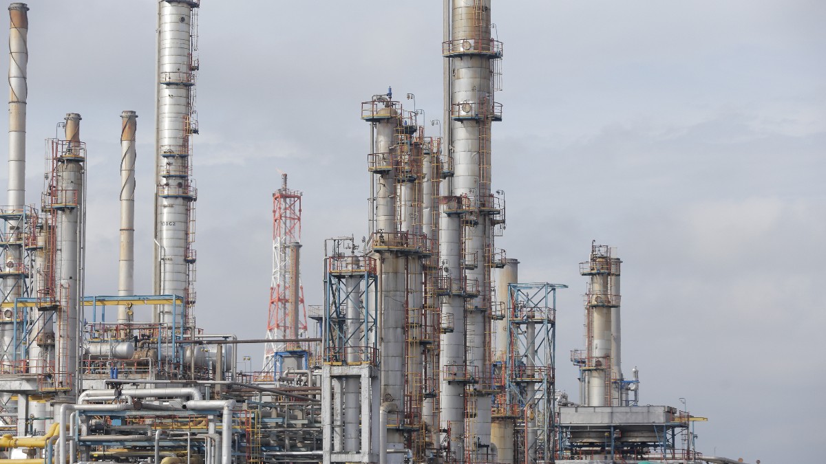 Imaginea surprinde un peisaj industrial vast, dominat de o rafinărie de petrol sau o uzină petrochimică, ilustrată de o rețea complexă de turnuri de distilare, conducte și structuri metalice. Instalația este fotografiată pe un fundal de cer albastru senin, cu o iluminare puternică, sugerând o zi de lucru obișnuită.