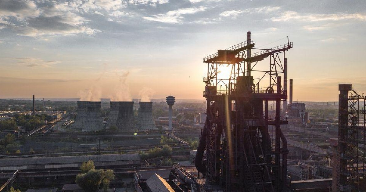 Imaginea aeriană surprinde un peisaj industrial vast, dominat în prim-plan de structura metalică masivă a unui furnal sau a unei cocserii, silueta fiind conturată de lumina puternică a apusului. În planul îndepărtat, se disting turnuri de răcire înalte, din care iese abur, și un turn de apă, confirmând că scena reprezintă Combinatul Siderurgic Liberty Galați.