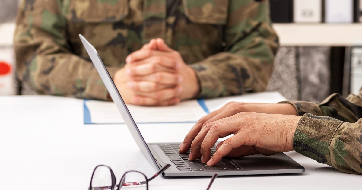 În imagine sunt prezentate în plan apropiat două persoane ce poartă uniforme militare de camuflaj așezate la o masă albă, una dintre ele tastând la un laptop lângă o pereche de ochelari, în timp ce cealaltă are mâinile împreunate în fundal.