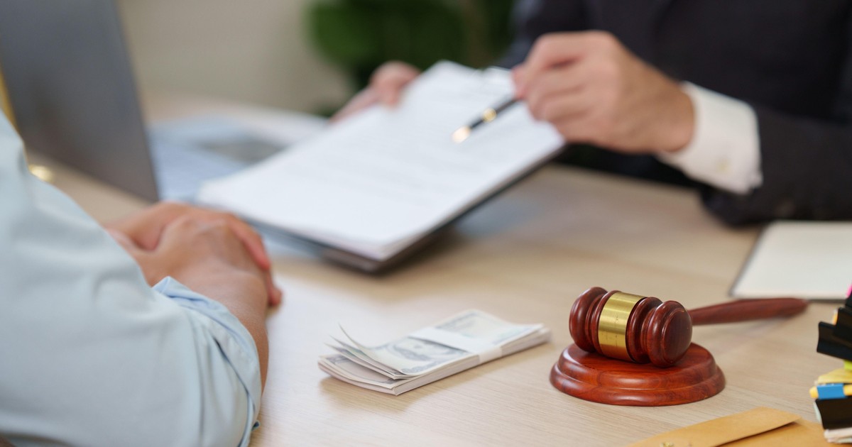A close-up of a wooden gavel and a stack of US dollar bills on a desk, with a blurred background of a lawyer presenting a contract to a client.,Image: 1116810451, License: Royalty-free, Restrictions: , Model Release: no