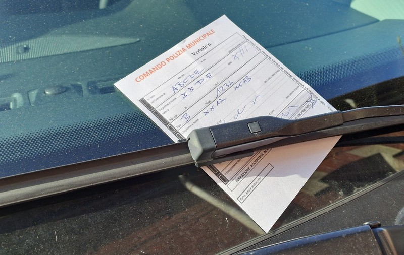 O înștiințare de amendă emisă de poliția locală din Italia, completată de mână pe un formular tipizat, fixată sub ștergătorul de parbriz al unui vehicul.