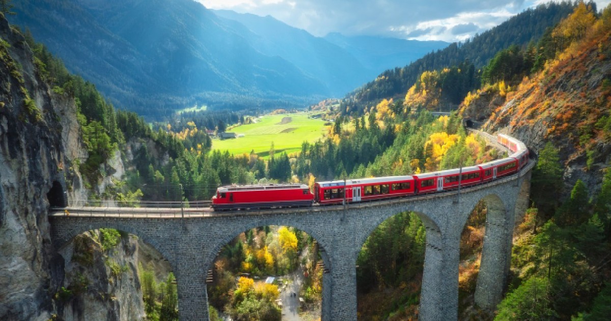 Vedere aeriană a trenului roșu modern pe viaductul Landwasser din munții alpini, pădure colorată la apus de soare în toamnă. Bernina Express, Elveția. Vedere de sus a trenului în Alpi, cale ferată, copaci verzi în toamnă