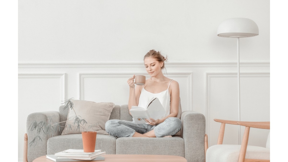 Femeie care sta in sufragerie pe o canapea de 3 persoane gri, se relaxeaza cu o cafea in mana si citind