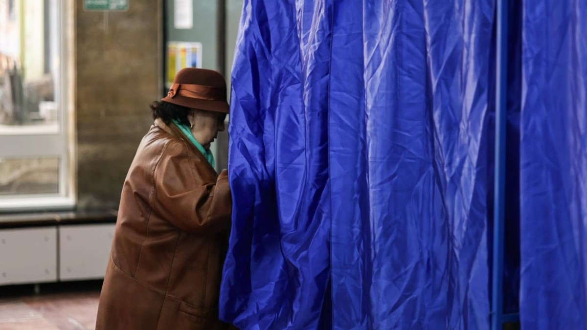 femeie în vârstă îmbrăcată într-un palton maro, cu o pălărie cu paglică maro, care intră într-o secție de votare