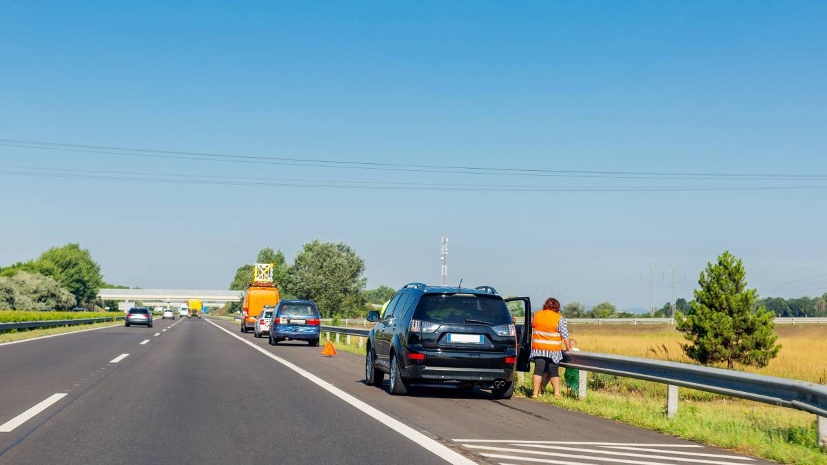 Mai multe mașini oprite pe banda de urgență de pe autostradă. Prima mașină este o mașină neagră, iar în dreapta ei este o femeie cu vestă reflectorizantă. În fața mașinii negre sunt alte două mașini oprite, care au triunghiul reflectorizant montat.