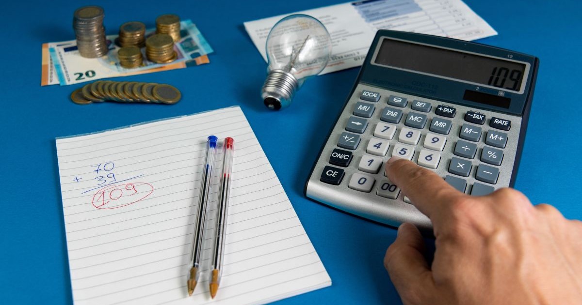 O factură de electricitate cu un bec, o mână calculează factura folosind un calculator și un caiet cu două pixuri și multe monede euro pe fundal albastru, iar sub ele este o hârtie cu un calcul și două pixuri, unul albastru și unul roșu