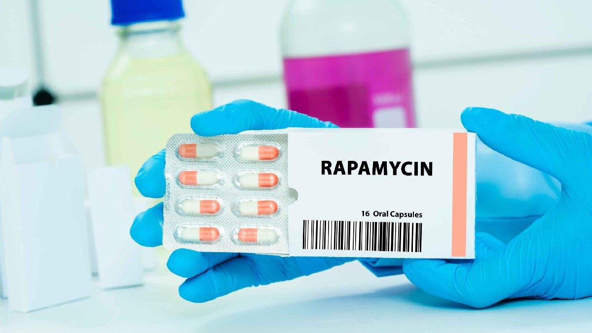 Imaginea prezintă de aproape o cutie de medicamente pe care este vizibil numele RAPAMYCIN și specificația "16 Oral Capsules", împreună cu un blister conținând capsule bicolore (alb și portocaliu). Mâini protejate de mănuși medicale albastre țin cutia, sugerând un mediu de laborator sau farmaceutic.