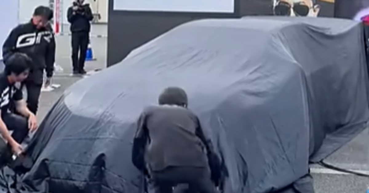 O mașină misterioasă, complet acoperită cu o husă neagră, este pregătită pentru dezvăluire în cadrul unui eveniment auto, în timp ce mai multe persoane se află în jurul ei. Silueta vehiculului sugerează un model sportiv sau de performanță, stârnind curiozitatea înaintea prezentării oficiale.