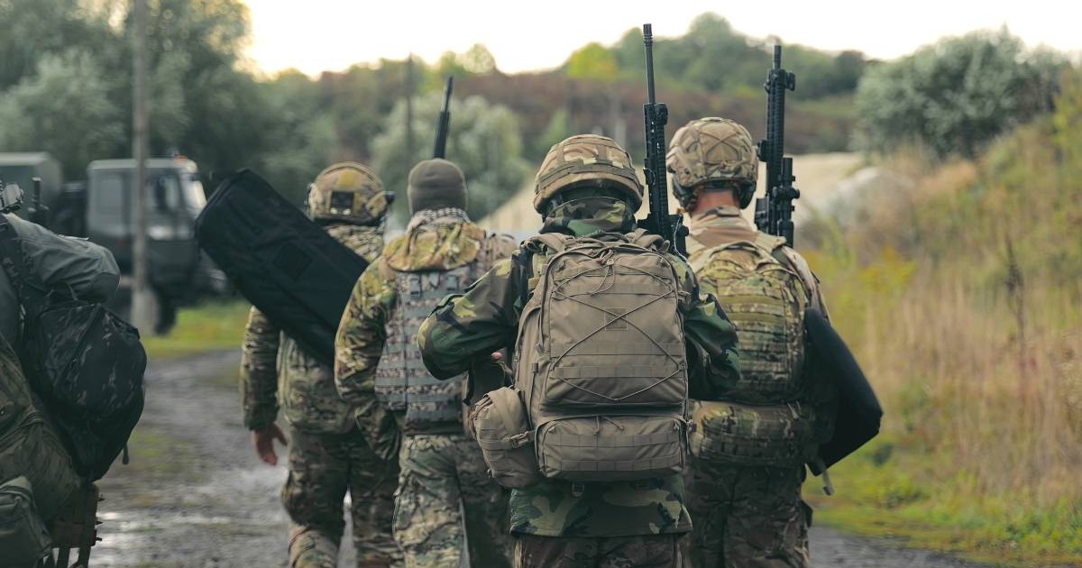 Un grup de militari în costume de camuflaj care merg cu puștile la spate pe stradă și se îndreaptă spre o cazară, toți privesc în jos