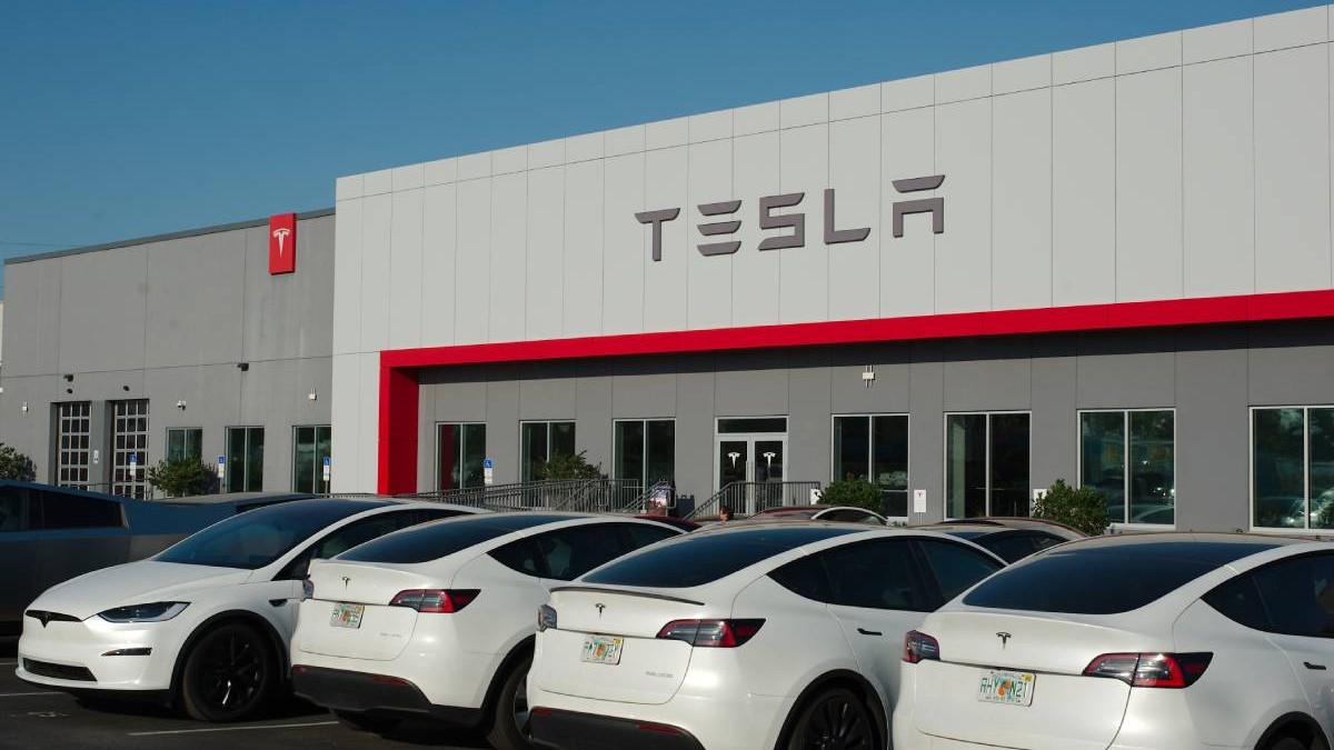 Exteriorul unei cladiri Tesla, un showroom sau un service auto. În fața clădirii sunt parcate mai multe vehicule Tesla albe, în special modele Model Y și Model X