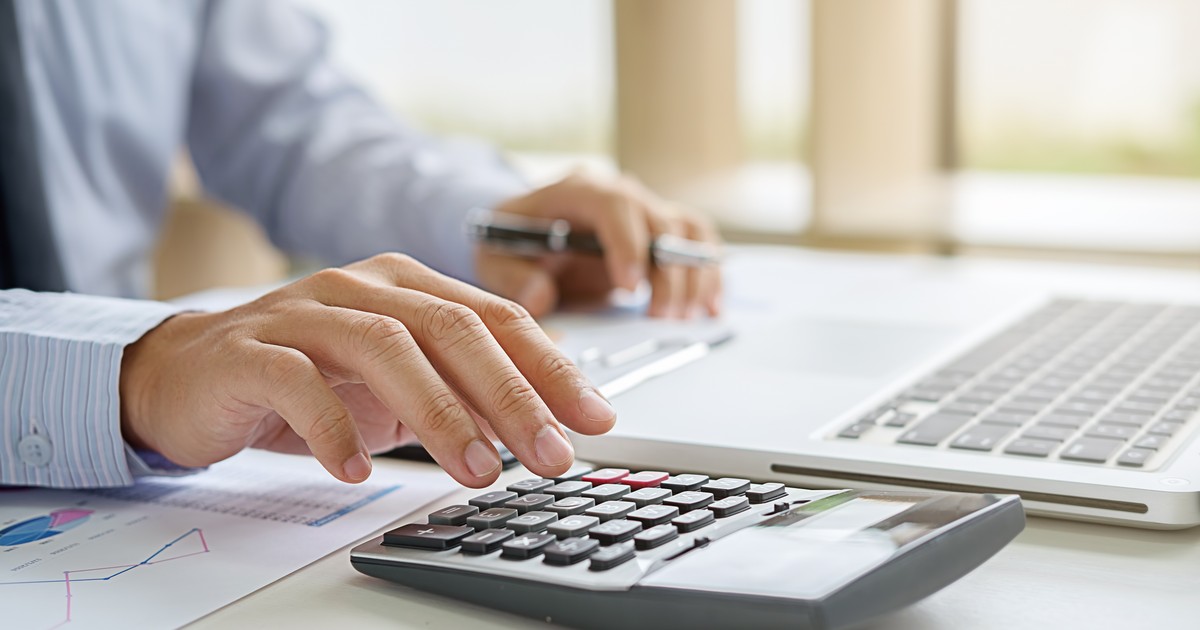 economist care calculează folosind un calculator, în timp ce sta cu o mana, în care ține un pix așezat între degete, peste un document printat pe o coală albă de hârtie și consultă datele disponibile pe ecranul unui laptop