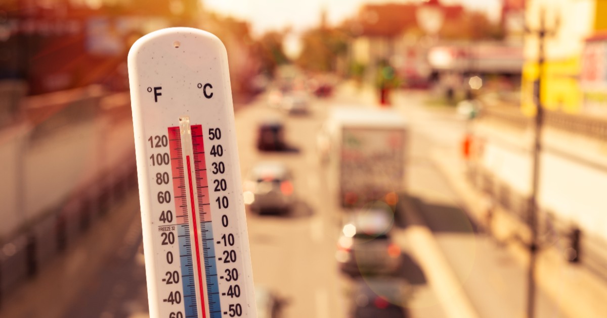 Imaginea prezintă un termometru alb care indică o temperatură ridicată de aproape 40°C, pe fundalul estompat al traficului rutier dintr-un oraș în plină caniculă.