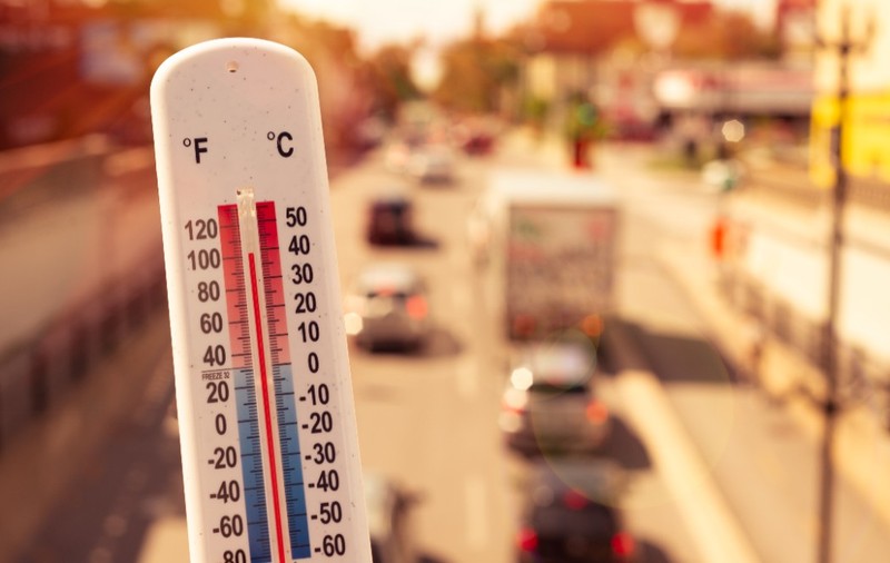 Prim-plan cu un termometru de exterior care indică o temperatură ridicată, pe fundalul unui drum urban însorit și aglomerat cu vehicule.