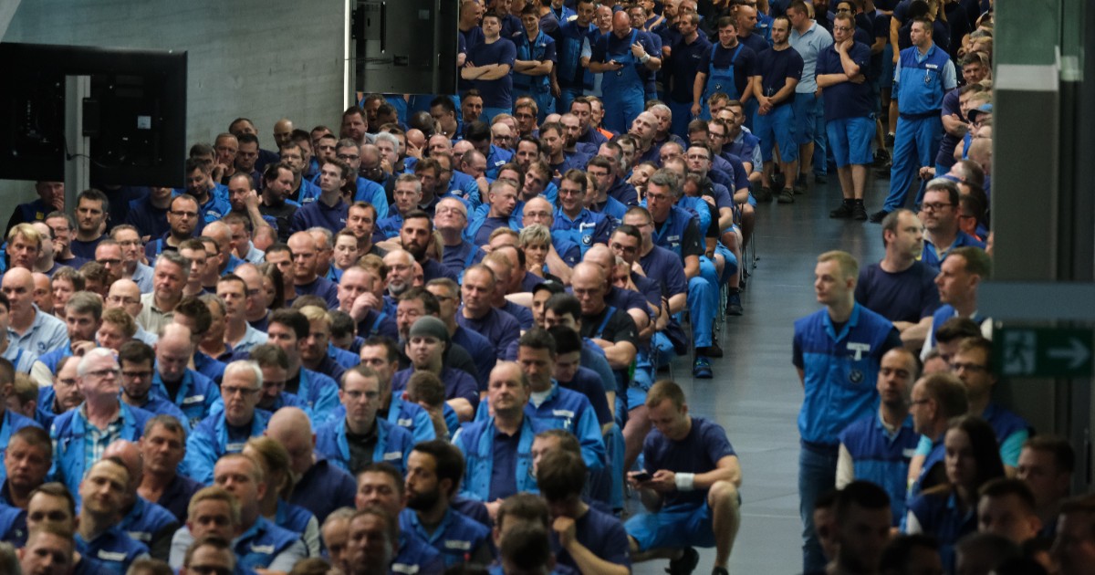 Imaginea prezintă un grup numeros de muncitori îmbrăcați în salopete albastre, adunați într-o sală mare a unei fabrici pentru o ședință sau o adunare generală.
