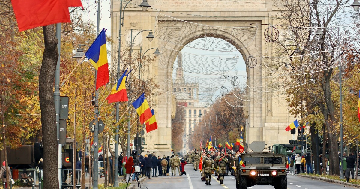 O paradă militară desfășurată pe stradă, cu Arcul de Triumf din București vizibil în fundal. Pe marginea străzii se văd steagurile României, iar în plan apropiat se văd militari și un vehicul militar.