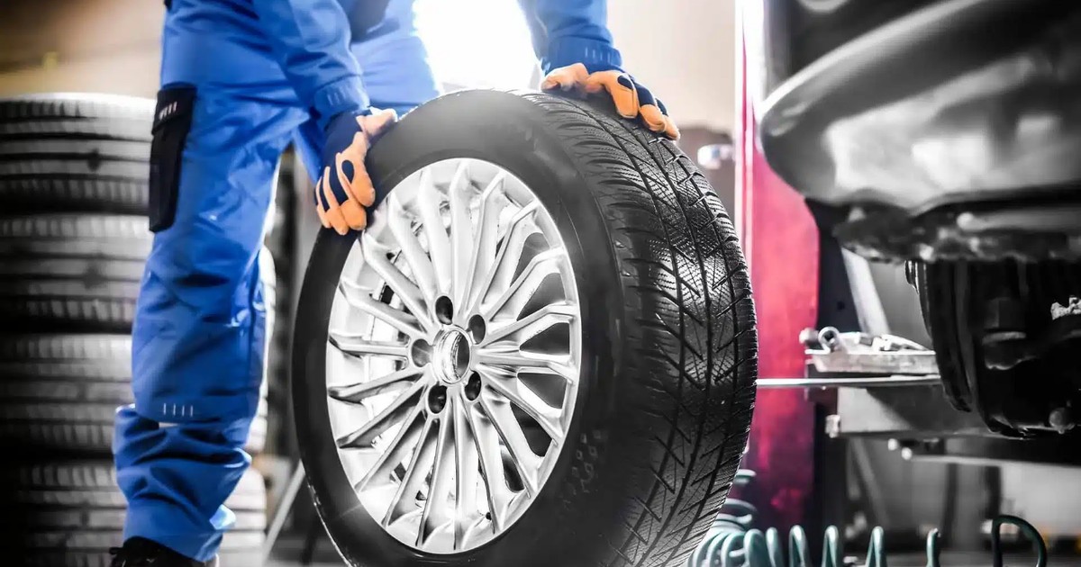 Imaginea surprinde un mecanic echipat cu o salopetă albastră și mănuși de protecție care sprijină cu ambele mâini o roată de mașină cu jantă argintie, pregătindu-se să o monteze pe un vehicul ridicat în cadrul unui atelier auto profesionist.