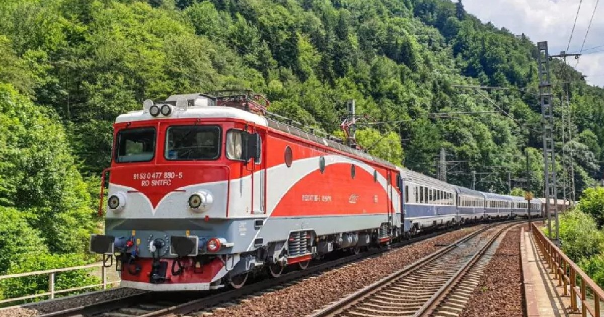 Un tren alb și roșu circulă pe lângă o pădure