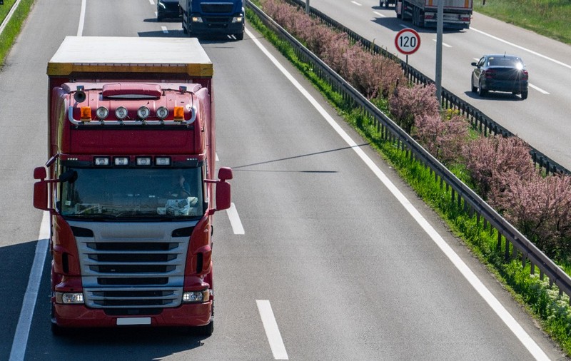 Un camion roșu rulând pe prima bandă a unei autostrăzi aglomerate, încadrat de alte autovehicule și un parapet median cu vegetație.