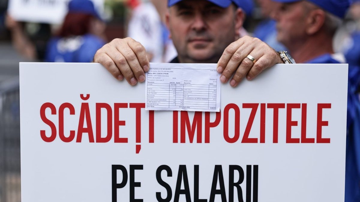 bărbat care poartă o șapcă albastră și haine albastre, ținând în mână o pancartă c text roșu și un fluturaș de salariu. Bărbatul poartă pe degetul inelar de la mâna stângă o verighetă groasă de aur. în spatele său se pot observa mai multe persoane, atât femei și bărbați, îmbrăcați în diverse articole de îmbrăcăminte albastre