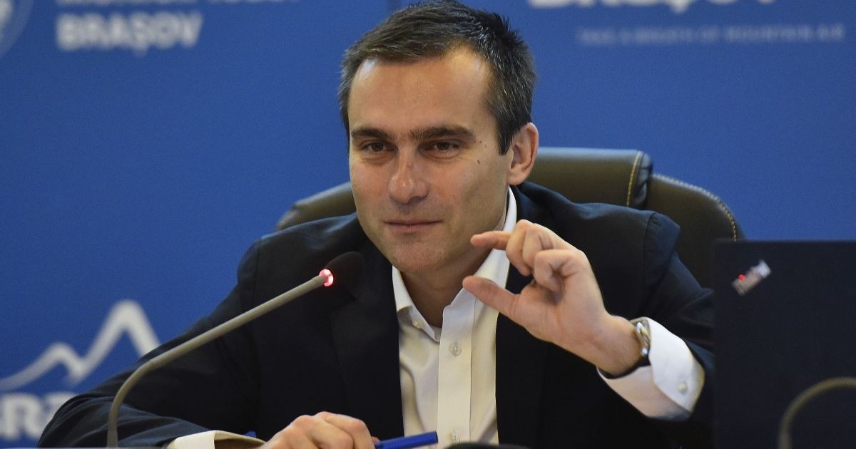 Allen Coliban, in sacou negru si camasa albă, surprins in timp ce vorbeste si face un semn cu mana stanga, 2022