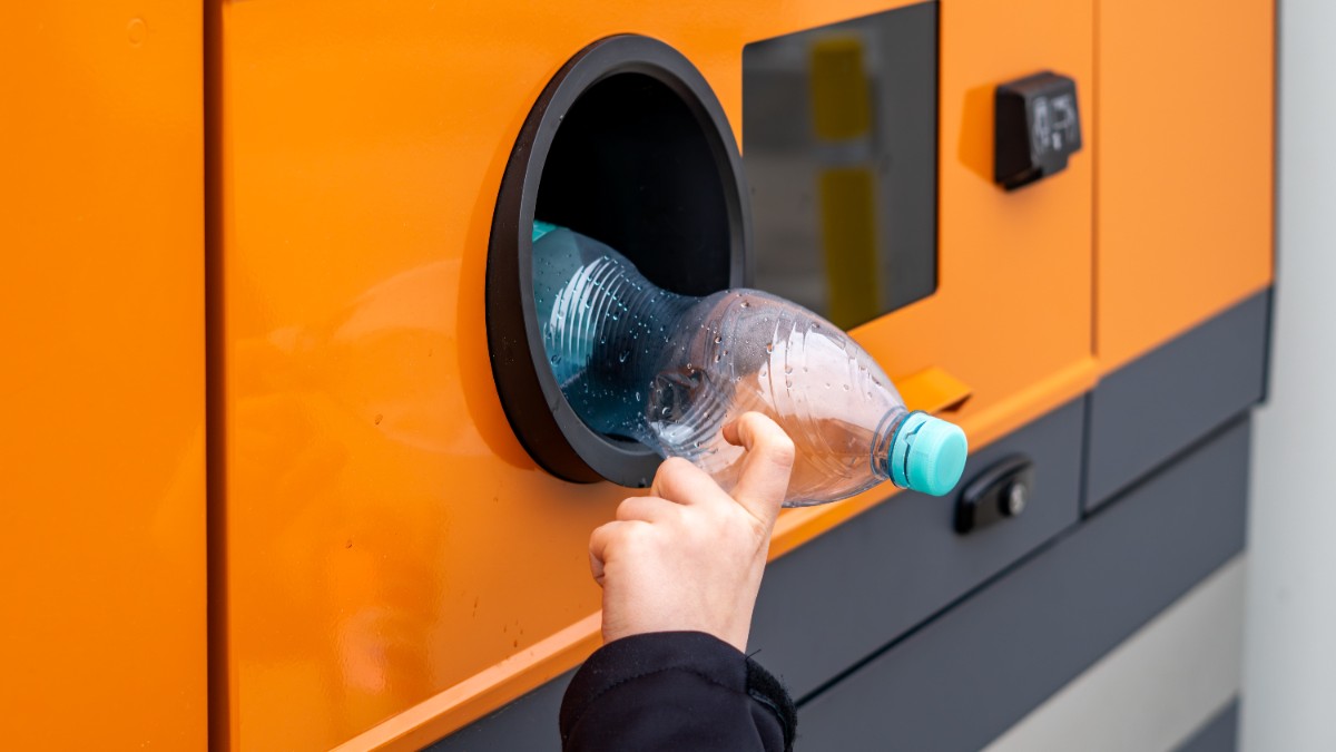 Imaginea prezintă o mână care introduce o sticlă goală de plastic într-un aparat de reciclare.