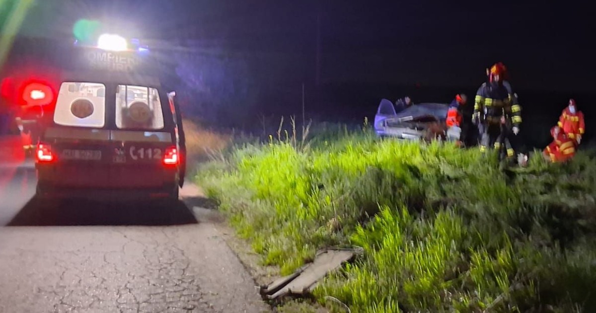 Imaginea surprinde intervenția echipajelor de urgență la locul unui accident rutier petrecut pe timp de noapte, pe marginea unui drum. În apropiere se observă o mașină avariată, iar pompierii și paramedicii acționează pentru gestionarea situației.