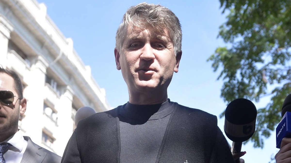 Cristian Pomohaci, îmbrăcat cu o bluză neagră și o vestă neagră, lângă o clădire, cu cerul albastru pe fundal
