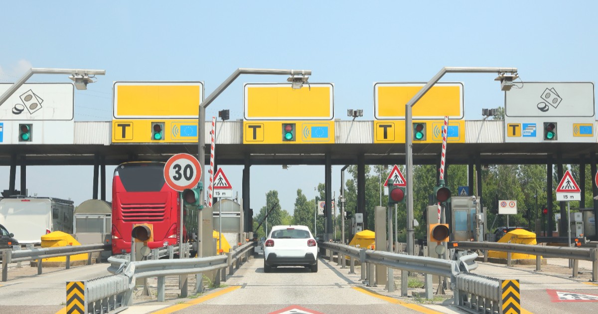 Imaginea prezintă o stație de taxare pe o autostradă, având mai multe benzi prevăzute cu bariere, semafoare și indicatoare rutiere, prin care trec un autoturism alb și un autocar roșu.