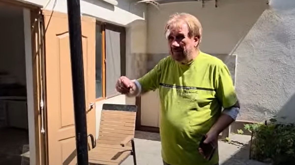 Gigel Slabu, membru AUR, in tricou verde, cu un bandaj la cap si unul la mana, tine mana dreapta ridicata si explica cum a fost atacat in propria curte, in spate se vede casa si un scaun de gradina