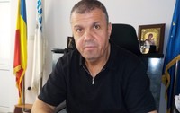 Imaginea îl prezintă pe Dănuț Buhăescu, îmbrăcat într-o bluză neagră, stând la biroul său încadrat de drapelul României și cel al Uniunii Europene.