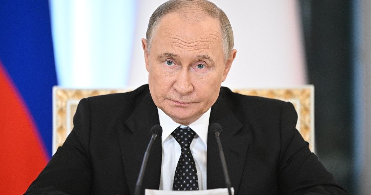 Prim-plan cu președintele Vladimir Putin, îmbrăcat într-un costum închis la culoare, are două microfoane în față și ține în mâini o bucată de hârtie, cu steagul Rusiei în fundal