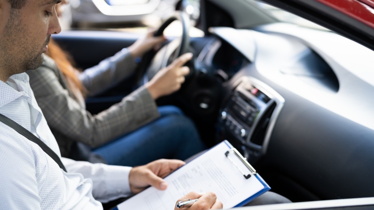 În imagine este ilustrat momentul examenului pentru permisul auto. O tânără se află la volan, iar examinatorul în partea dreaptă.